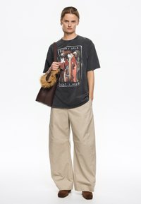 Camiseta gráfica de gran tamaño en gris oscuro con un diseño colorido, combinada con pantalones de pierna ancha en crema y un bolso marrón con acento de piel sintética.