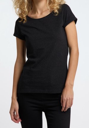 Mujer con cabello rubio rizado que lleva una camiseta negra de manga corta con finas rayas horizontales y pantalones negros sobre un fondo liso claro.