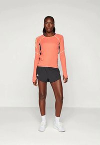 Cămașă sport cu mânecă lungă de coral, cu accente în nuanță de albastru închis și cusături deschise, purtată cu pantaloni scurți negri. Modelul poartă adidași albi și șosete până la gleznă.