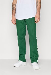 Zöld, fehér átlós csíkokkal díszített melegítőnadrágot és fehér sportcipőt viselő férfi áll sík háttér előtt.