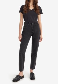 Jeans de mezclilla negros de cintura alta con pierna ajustada, que incluyen un cierre de botones, cinco bolsillos y un dobladillo deshilachado; combinados con zuecos negros sin talón.