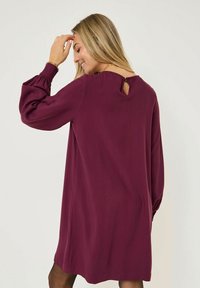 Donna con lunghi capelli biondi che indossa un vestito a maniche lunghe color prugna, si sta toccando la fronte e sorridendo, rivolta leggermente di lato.