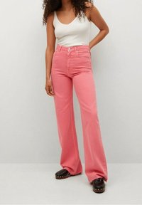 Jean rose clair taille haute avec jambe évasée, doté d'une fermeture à bouton devant et de passants pour ceinture, porté avec un débardeur côtelé blanc et des sabots.