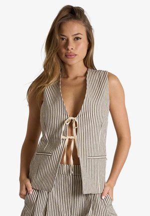 Donna che indossa un gilet smanicato beige e bianco a righe verticali, legato con due fiocchi sul davanti e pantaloni a vita alta abbinati.