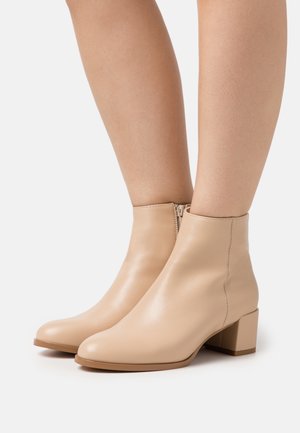 Bottes beige à cheville avec des talons blocs et des zips latéraux, portées sur des jambes nues devant un fond blanc uni.
