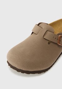 Sabot en suède beige avec un bout arrondi, doté de deux sangles ajustables avec une boucle en métal, d'une texture lisse et d'une semelle marron texturée.