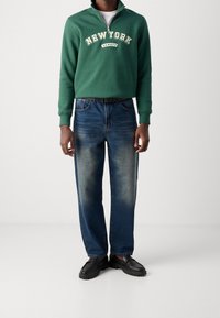 Zelená mikina se zipem u krku s nápisem "NEW YORK", doplněná modrými džíny s rovnými nohavicemi. Černé nazouvací boty dokončují outfit.