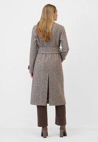 Lång brun texturerad kappa med bälte i midjan, ryggslits och ljusprickigt mönster, bärs över bruna vida byxor och klackstövlar.