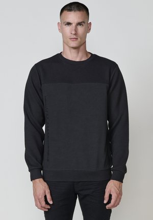 Sudadera negra de manga larga con tejido texturizado, panel frontal liso, puños y dobladillo de canalé. Presenta un detalle de logo vertical en el lateral.