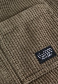 Tissu en velours côtelé marron avec cannelures verticales ; présente une étiquette noire avec du texte blanc. Textures et matériau visibles en gros plan.