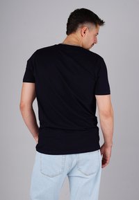 Mann mit kurzen dunklen Haaren, der ein schwarzes T-Shirt und hellblaue Jeans trägt, steht mit dem Rücken zu einem schlichten grauen Hintergrund.