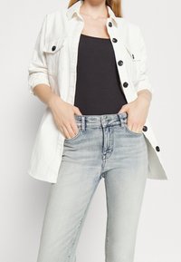 Camisa de mezclilla blanca con botones negros, llevada sobre una camiseta sin mangas negra, combinada con jeans ajustados de un azul claro con una textura descolorida.