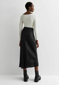 Larga falda negra de satén combinada con una blusa blanca de manga larga ajustada; presenta una textura suave y un ligero brillo. Botas negras gruesas completan el look.