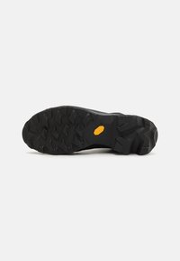 Semelle de chaussure de sport noire avec une surface rugueuse et texturée, dotée de crampons proéminents pour une meilleure traction. Logo Vibram visible en jaune.