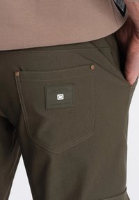Mörkgröna byxor i en texturerad tyg, med en rektangulär logotyp patch på bakfickan och försedda med två kopparfärgade nitar.