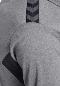 Zbliżenie na szarą tkaninę o fakturze z czarnymi detalami wentylacyjnymi i wzorem w jodełkę na obszarze ramienia odzieży.