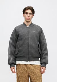 Tânăr cu păr scurt brunet, purtând o jachetă din fleece gri cu fermoar și pantaloni texturați maro, stând în fața unui fundal alb simplu.