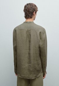 A person with short, wavy hair viewed from the back wears an olive-green, long-sleeved shirt in a neutral-toned setting.