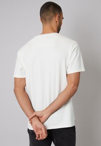 Weißes T-Shirt mit kurzen Ärmeln aus strukturiertem Stoff, runder Halsausschnitt und einem kleinen quadratischen Akzent auf dem oberen Rücken. Das Modell ist abgewandt.