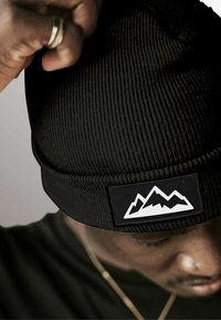 Black knit beanie with a textured pattern. Features a rectangular patch displaying a white mountain graphic. Close-up of fit.