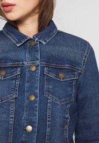 Denim jacket in dark blue with a collar, front button closure, and two chest pockets featuring metallic buttons and contrast stitching.