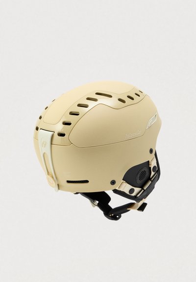 Casco da sci beige con fessure di ventilazione, cinturino regolabile e dettagli neri. Superficie in plastica liscia; presenta un gancio laterale per una vestibilità sicura.