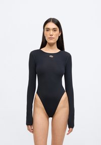 Μαύρο κορμάκι με μακριά μανίκια και ψηλή κοπή στο πόδι, λεία υφή και μικρή μεταλλική λεπτομέρεια με λογότυπο στο στήθος. Σχεδίαση που εφαρμόζει στο σώμα.