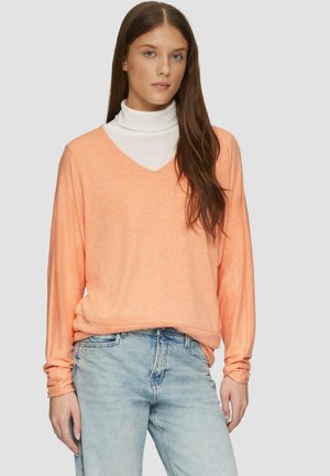 V-Ausschnitt, langärmliges Oberteil in Hellorange, aus weichem Stoff, über einem weißen Rollkragenpullover getragen, kombiniert mit hellblauen Jeans.