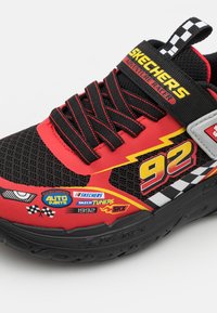 Zapatilla roja y negra con parte superior de malla, presenta gráficos brillantes, diseño de rayo y múltiples logotipos. Incluye cordones y correa negra.