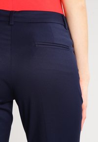 Marinblå byxor med en mjuk textur, skräddarsydd passform och en bakficka. Midjebandet är platt och kompletteras av en röd topp.