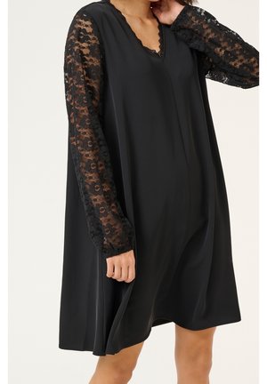 Femme portant une robe noire ample avec des manches longues en dentelle florale transparente et un décolleté en V, main touchant doucement le cou.