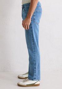 Osoba w jasnoniebieskich dżinsach prostych i białych niskich trampkach z gumową podeszwą stoi przy teksturowanej białej ścianie.