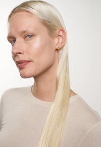 Ljusbeige, figursydd långärmad skjorta, texturerad. Lång rak platinablond hår. Guld hoop-örhänge i vänstra örat. Naturlig hudton, subtil glans.