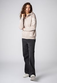 Beige zip-up hoodie met witte gedrukte tekst op de linkerarm, voorzien van een trekkoordcapuchon en zijvakken. In combinatie met donkere jeans en sneakers.