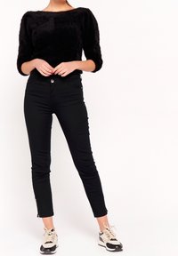 Mujer vestida con un top negro de felpa, pantalones ajustados negros con cremalleras en los tobillos y zapatillas de deporte beige y negras, de pie sobre un fondo blanco.