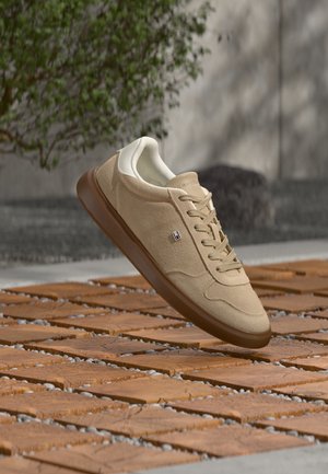 Baskets en daim beige avec semelle marron et petit logo sur le côté, flottant au-dessus de carreaux de bois décorés en extérieur près d'un mur et de verdure.
