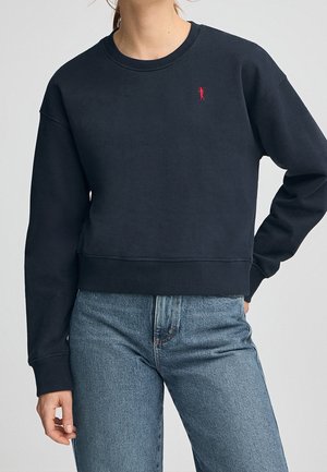 Kobieta w czarnej bluzie z okrągłym dekoltem z małym czerwonym logo golfisty i niebieskich jeansach z wysokim stanem, stojąca z jedną ręką na biodrze.