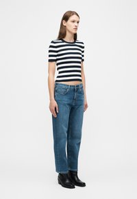 Γυναίκα που φοράει μπλούζα με μαύρες και άσπρες ριγές και κοντά μανίκια, τζιν μπλε με ίσια πόδια και μαύρα μποτάκια αστραγάλου, στέκεται μπροστά σε ένα απλό φόντο.