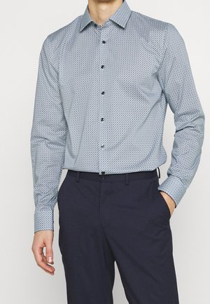 Mann trägt hellblaues, gemustertes Langarmhemd, in dunkelblaue Hose gesteckt, mit einer Hand in der Tasche.