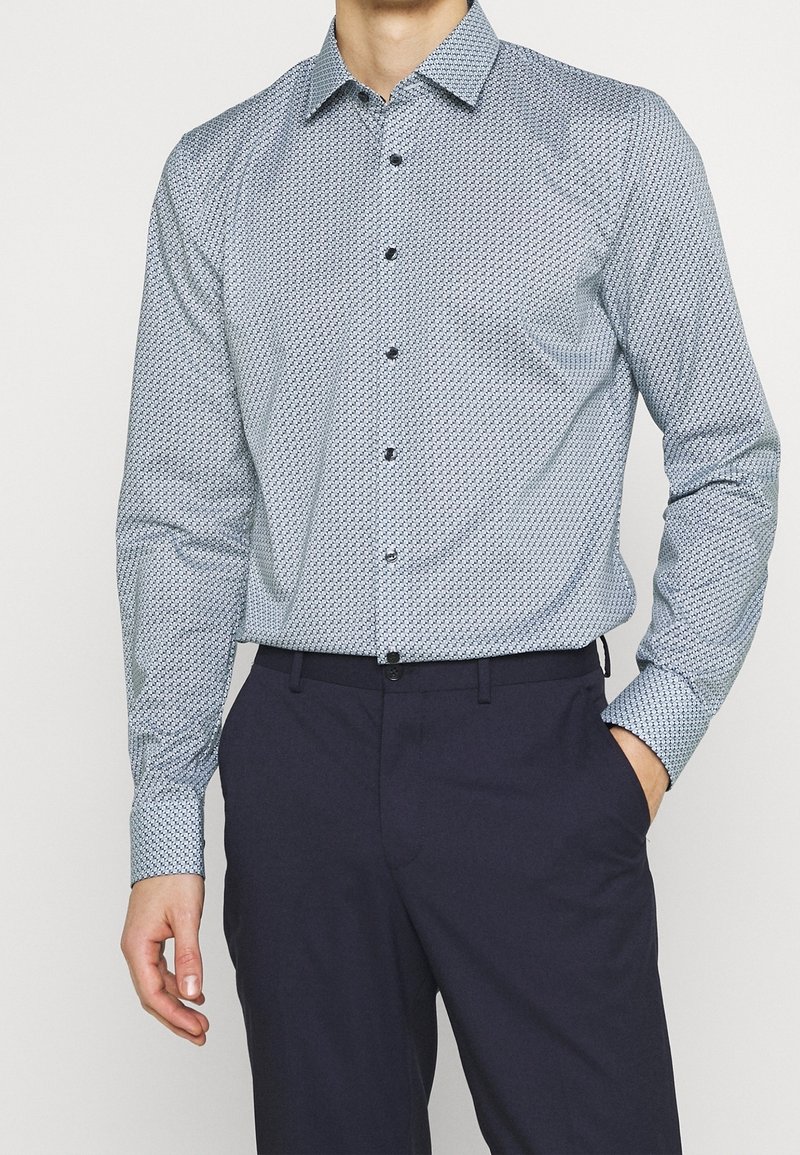 Homme portant une chemise à manches longues bleu clair à motifs, rentrée dans un pantalon bleu marine foncé, une main dans la poche.