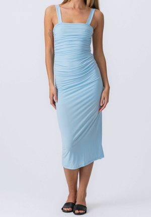 Frau trägt ein hellblaues, gerafftes, ärmelloses Midikleid mit breiten Trägern und schwarze offene Slip-On-Sandalen, steht vor einem schlichten Hintergrund.