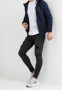 Sudadera con cremallera azul marino sobre una camiseta blanca, combinada con jeans ajustados negros rasgados y zapatillas blancas con detalles en gris.