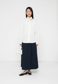 Camicia bianca con un dettaglio al collo legato, abbinata a una gonna maxi plissettata blu navy. Accessoriata con una piccola borsa bianca e ballerine nere.