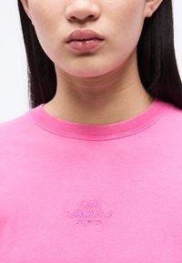 Κοντινό πλάνο ενός ατόμου που φοράει φωτεινό ροζ πουλόβερ με στρογγυλή λαιμόκοψη, στο οποίο είναι κεντημένη η φράση "MADS NØRGAARD COPENHAGEN" με νήμα που ταιριάζει στο ίδιο ροζ χρώμα.