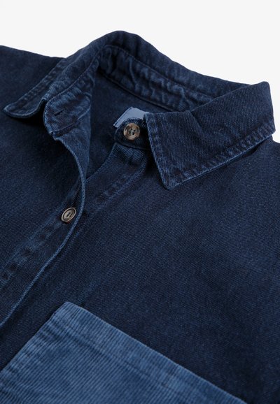 Chemise en denim bleu foncé avec un col plié, des boutons marron et une poche poitrine en velours côtelé bleu clair.