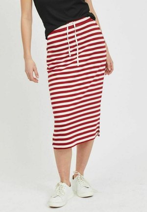 Kvinde iført en rød- og hvidstribet midi-nederdel med snøre, sort top og hvide sneakers mod en ensfarvet baggrund.