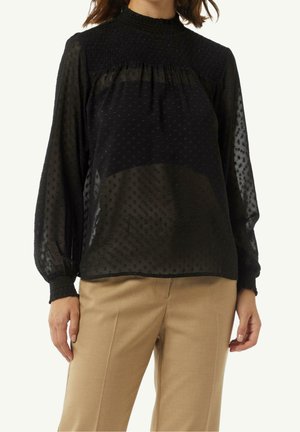 Femme portant un chemisier noir transparent à manches longues avec des pois texturés et un col et des poignets smockés, associé à un pantalon beige.