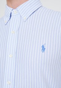Jasnoniebieska koszula na guziki w paski, wykonana z fakturowanej tkaniny. Posiada białe guziki oraz małe niebieskie haftowane logo na piersi.