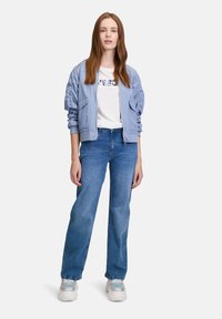 Hellblaue Bomberjacke mit gerippten Bündchen über einem weißen T-Shirt mit strukturiertem Druck, kombiniert mit blauen geraden Jeans und klobigen Sneakers.
