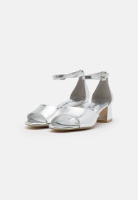 Sølvmetallic sandaler med åpen tå-design, ankelstropp og lav blokkheel. Glatt tekstur med en blank finish.